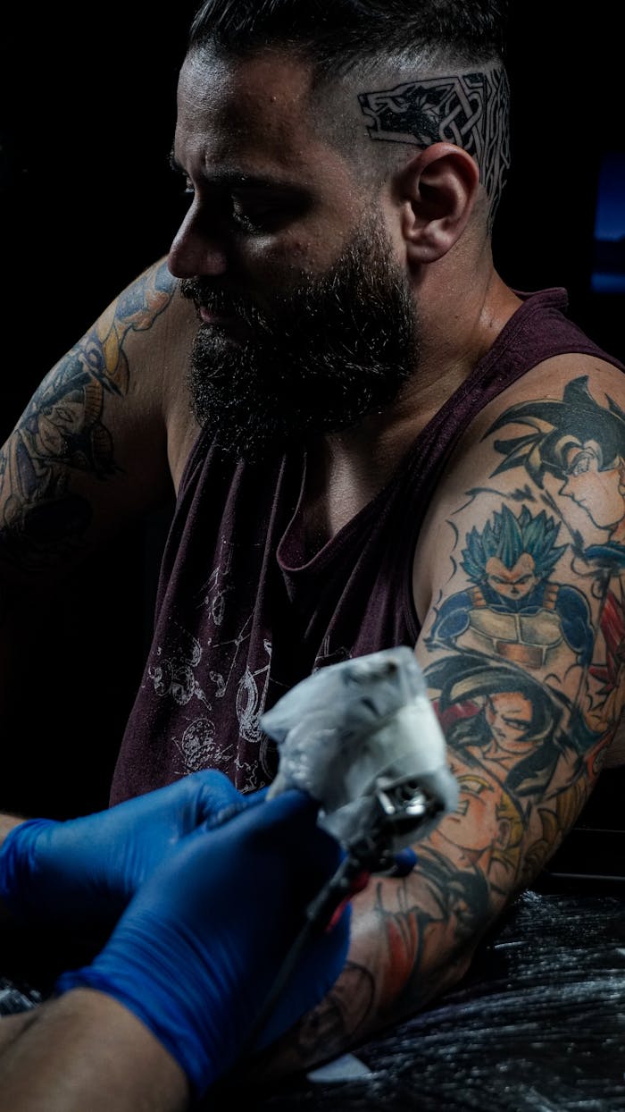 gallery-3 Bearded man in tank top receiving a vibrant arm tattoo in a studio setting.