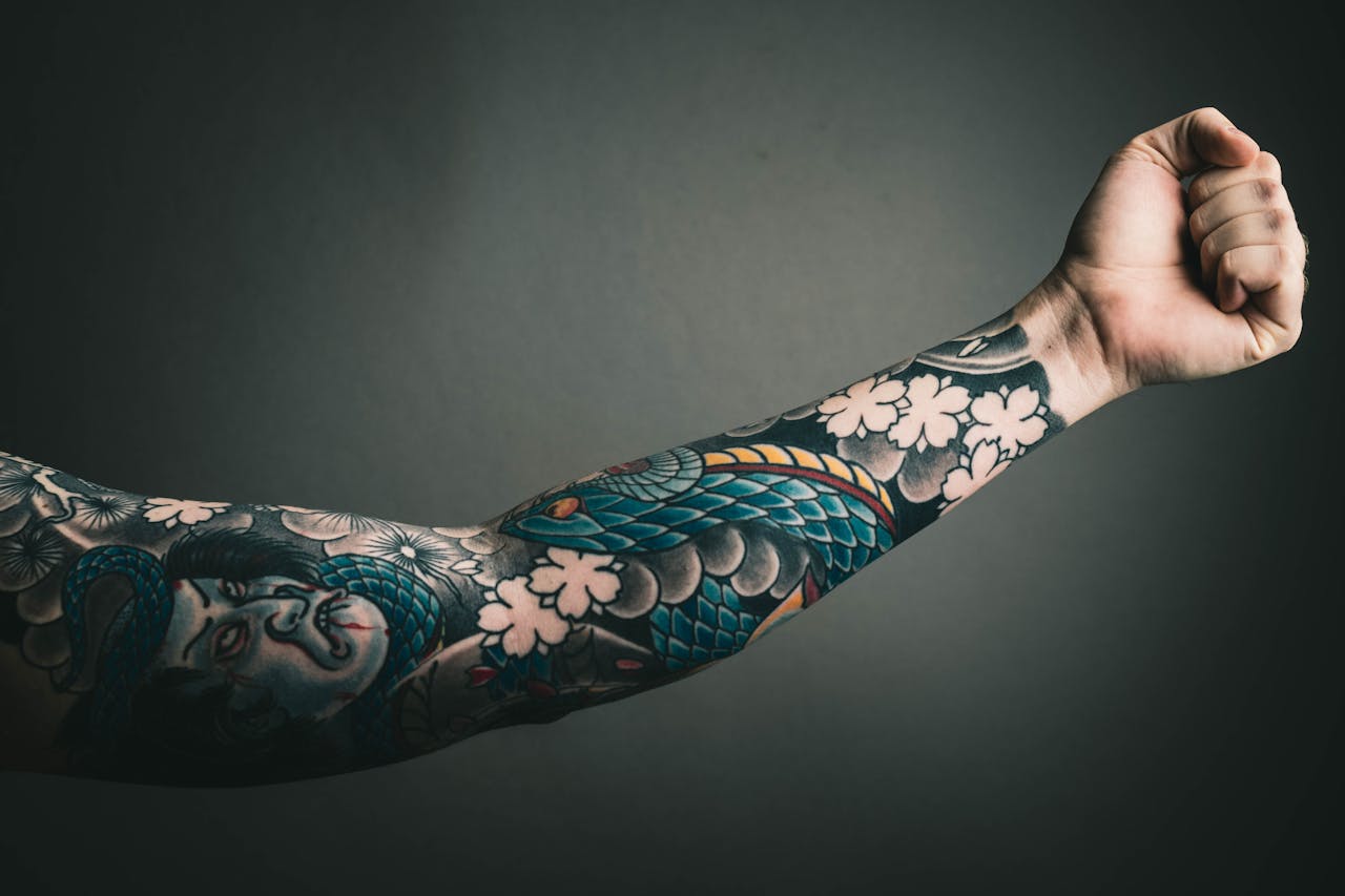 about-01 A close-up of a tattooed arm with a colorful sleeve design, featuring floral and artistic elements.