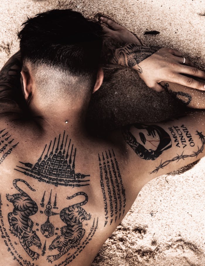 our-services-3 A tattooed man with intricate designs lying on a sandy beach, showcasing body art.